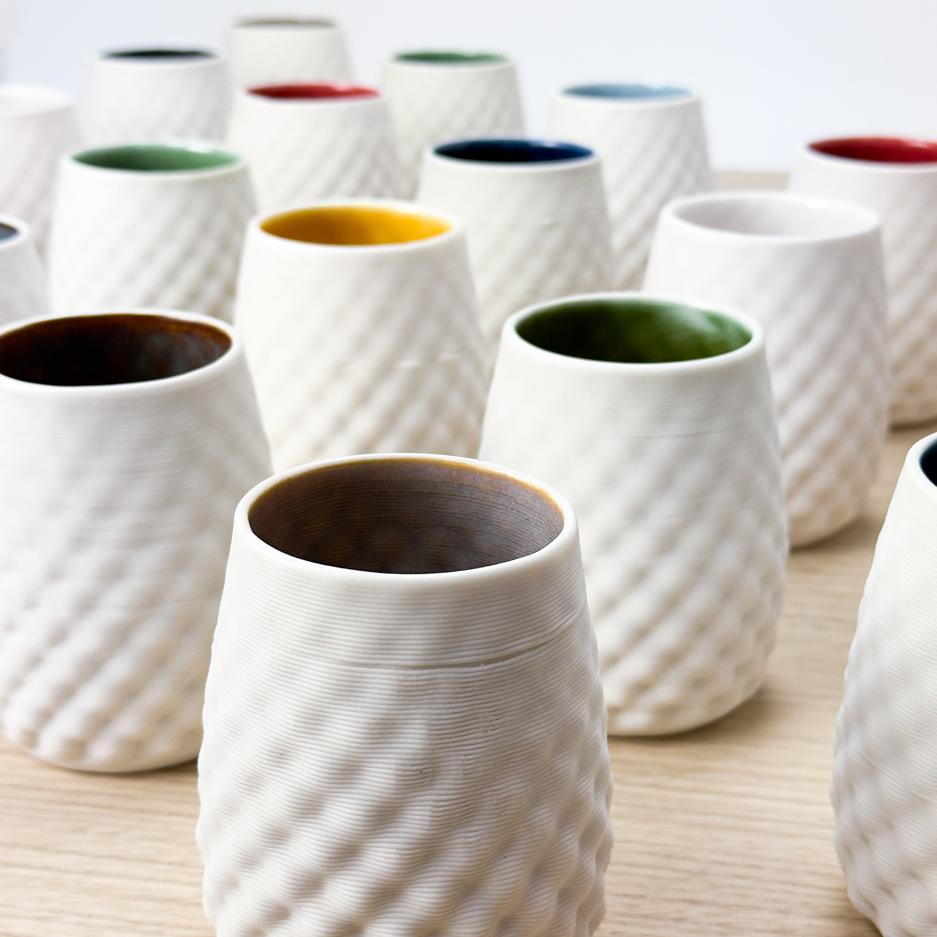 Set of white textured 3D printed ceramic cups with colorful interior glazes on a light wooden surface
