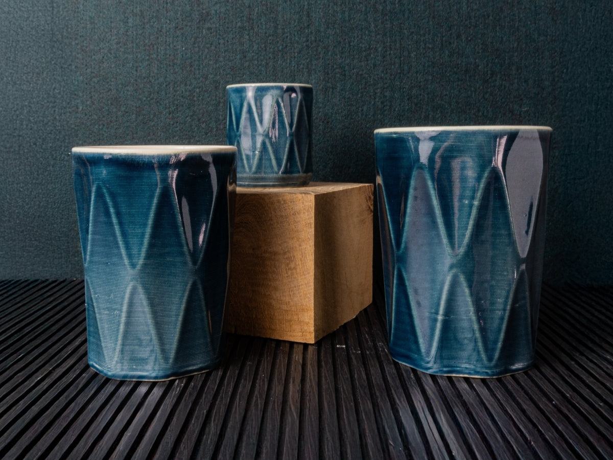 Three blue 3D printed ceramic cups with a diamond pattern on a dark surface against a dark background